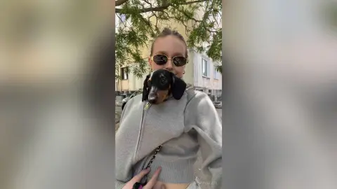 BonnyWalace wears sunglasses and a grey zip-up hoodie while holding a small dachshund dog outdoors near some trees.