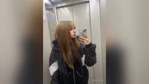 Young woman with long brown hair taking a mirror selfie in an elevator wearing a black and grey jacket.