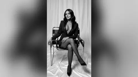 Elegant woman in a black blazer and stockings sitting on a chair in a studio setting with a sophisticated mood.