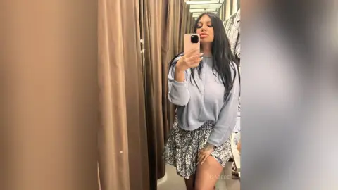 Young woman in a light blue sweater and floral skirt taking a mirror selfie in a clothing store fitting room.