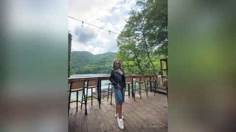 SelenaRusse stands on a wooden deck overlooking a lake, wearing a black leather jacket and denim shorts.