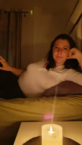 Zemer_secret51 lies on a bed with dark hair, wearing a white long-sleeve top, resting her head on her hand near a lit candle.