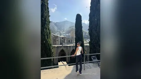 AnaMathers stands on a stone balcony wearing a white tank top and black pants, framed by tall cypress trees and a mountain view.