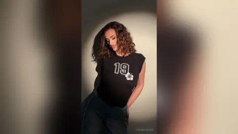 Smiling woman in a black graphic t-shirt posing against a neutral studio background with warm lighting.