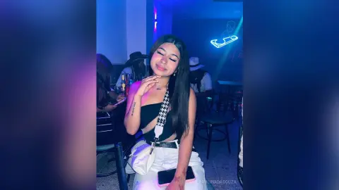 Young woman in a black crop top and white pants smiling in a moody, blue-lit nightclub setting.