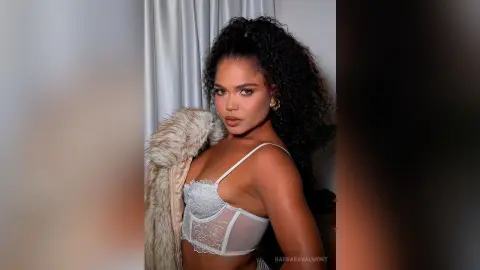 Confident woman in white lace lingerie and a beige faux fur coat posing against a neutral backdrop.