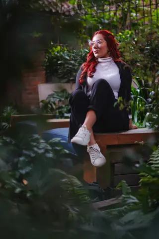 Red-haired woman in a white turtleneck and black trousers sitting peacefully in a lush green garden.