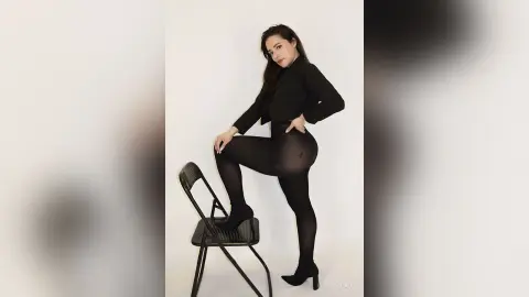 Confident woman in a black blazer and tights posing on a chair against a white studio background.