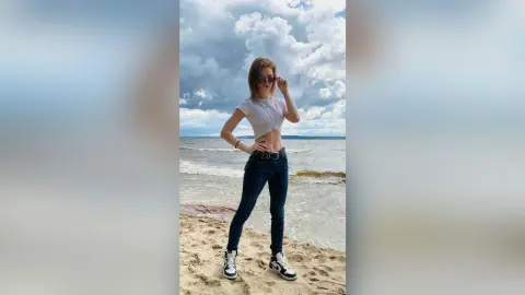 Young woman in white crop top and dark jeans posing on a sunny beach under a cloudy sky.