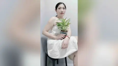 LouiseAnna sits on a black stool wearing a white strapless towel wrap, holding a small green potted plant against her chest.