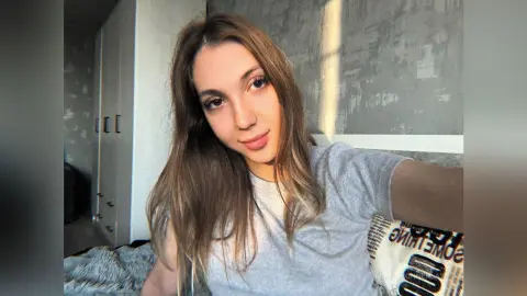 Young woman in a grey t-shirt relaxing in a sunlit bedroom with a cozy, serene mood.