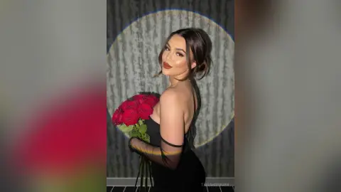 Elegant woman in a black dress holding red roses against a textured grey backdrop in moody lighting.