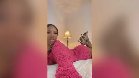 Smiling Black woman in cozy pink fuzzy loungewear relaxing on a bed in a warm, soft-lit bedroom.