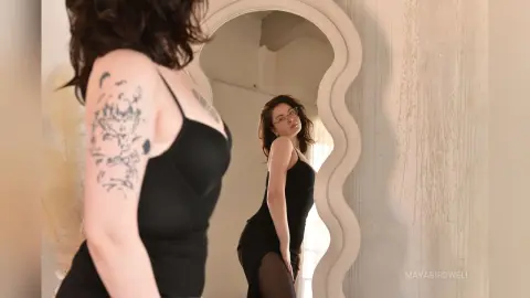 Woman with arm tattoos in a black slip dress looking into a wavy mirror in a bright, minimalist room.