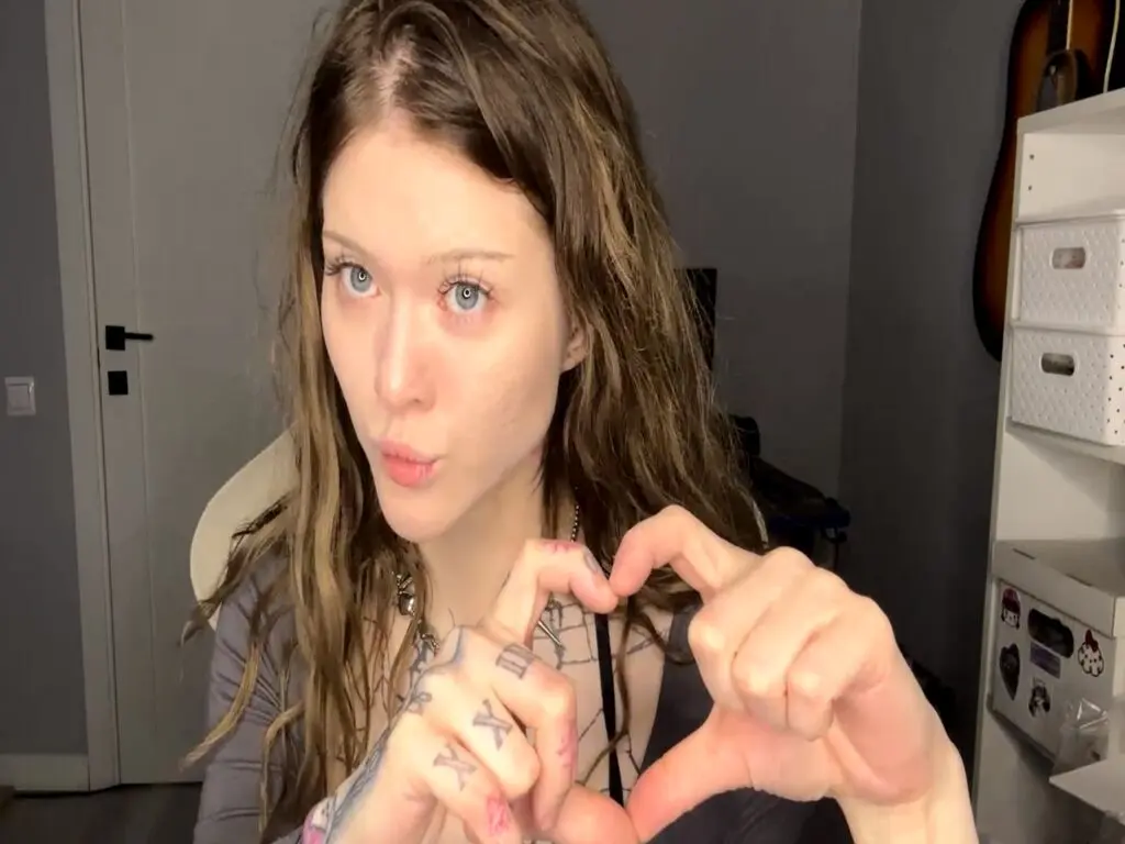 MaryLico makes a pouting face while making a heart shape with her hands, showing her long wavy hair and tattoos on her fingers.