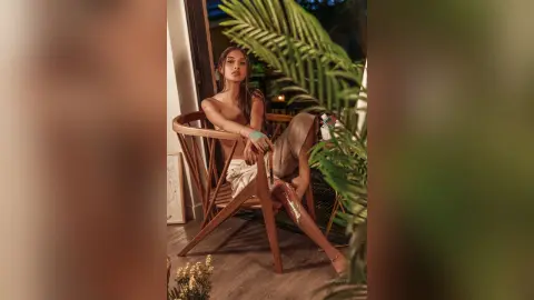 LucyMartin sits on a wooden chair wearing a light tan bra and matching bottoms, posing against a backdrop of green palm leaves.
