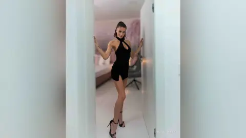 AnaMathers stands between two white doors wearing a short black halter-neck dress and black high-heeled sandals.