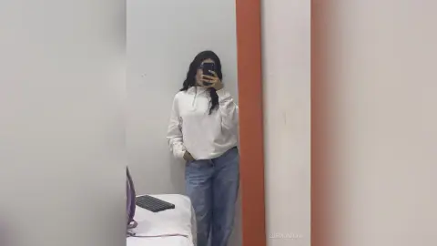 ZoeMonrow poses in a white hoodie and blue jeans, standing in a room with a white wall and a bed or massage table nearby.