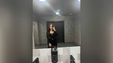Young woman in a black long-sleeve top and trousers taking a mirror selfie in a modern bathroom.