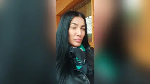 Smiling woman with long black hair wearing a black jacket and green scarf in a bright indoor setting.