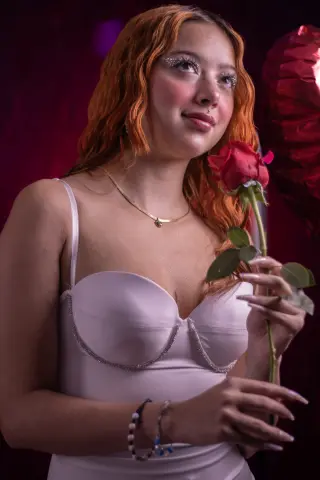 Young woman with red hair in a white slip dress holding a red rose in a moody, romantic setting.