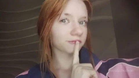 MozelleRozycki has red hair and holds a finger to her lips in a quiet pose against a dark, wavy background.