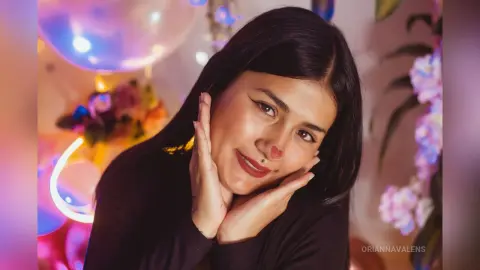 Smiling woman in a black top posing with hands on face in a dreamy, colorful room with soft lighting.