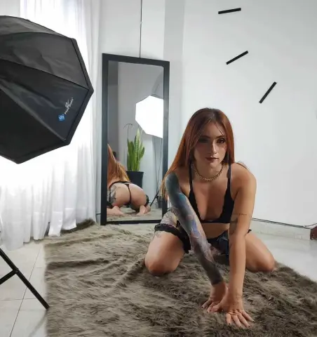 Tattooed woman in black bikini sitting on a shag rug in a bright photo studio setting.