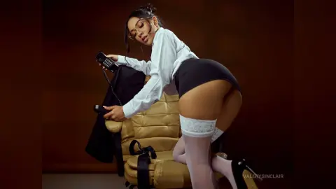 ValerySinclair wears a white shirt and black skirt, posing on a chair while holding a telephone.