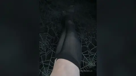 Close-up of legs in sheer black tights against a dark, moody background of intricate spider webs.
