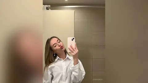 Young woman in a white button-down shirt taking a mirror selfie in a modern bathroom.