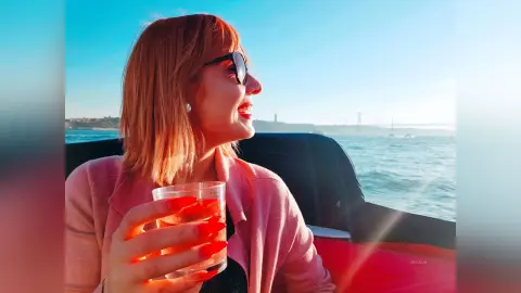 AryaSin smiles while wearing sunglasses and a pink cardigan, holding a clear glass of red liquid outdoors near the ocean.