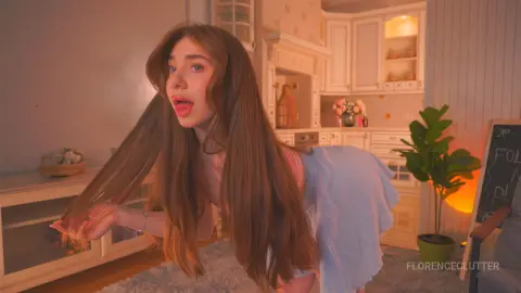 Young woman with long brown hair in a pink top posing in a cozy, warm-lit aesthetic kitchen setting.