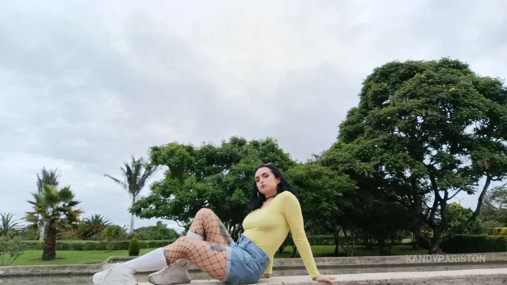 KandyPariston — Woman in a yellow long-sleeve top and denim shorts sitting outdoors in a lush green park under a cloudy sky.