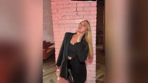 Blonde woman in a black blazer and crop top posing against a pink brick wall in a moody, stylish interior.