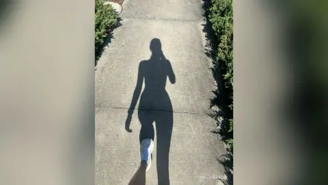 ChristineDavis casts a long shadow of her body on a concrete sidewalk, showing her silhouette and legs in white sneakers.