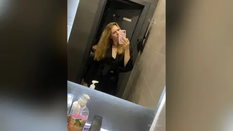 Blonde woman in a black outfit taking a mirror selfie in a modern bathroom setting with a moody vibe.