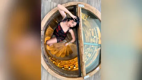 Fashionable woman in a black patterned dress relaxing in a circular wooden window nook with warm lighting.