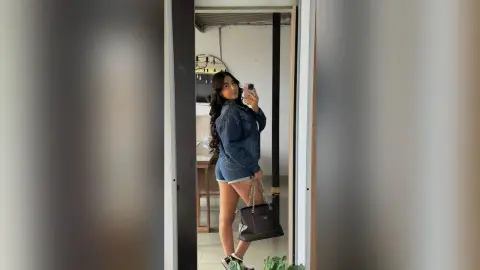 KimberlyIsaza poses in a denim shirt and denim shorts, holding a phone for a mirror selfie while carrying a black handbag.