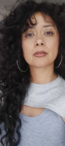 Woman with voluminous curly dark hair wearing a grey casual top in a soft, natural light setting.