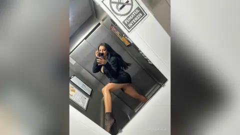 NicoleBella poses in a black dress and black stockings while standing in an elevator.