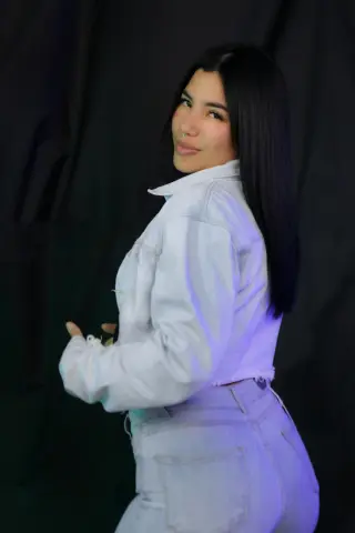 channeljonson — Smiling young woman with long dark hair wearing a light denim jacket and jeans in a studio setting.