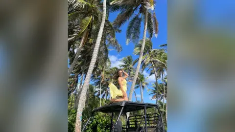 Cheerful woman in a yellow bikini relaxing on a vehicle roof under tropical palm trees on a sunny day.