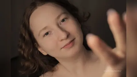 Close-up portrait of a young woman with wavy hair looking thoughtfully into the camera in soft, warm lighting.