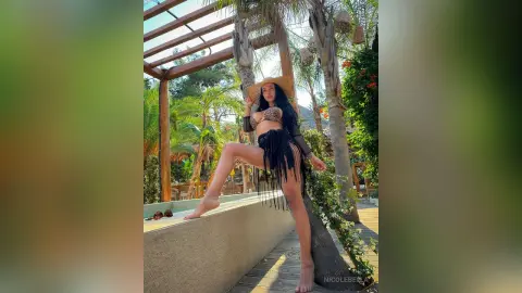NicoleBella poses outdoors in a black fringe bikini and straw hat, standing next to a stone ledge surrounded by palm trees.