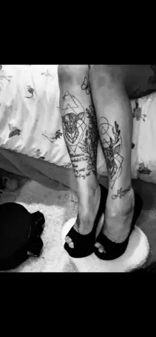 Close-up of tattooed legs wearing black high heel sandals resting on a white surface in a cozy indoor setting.