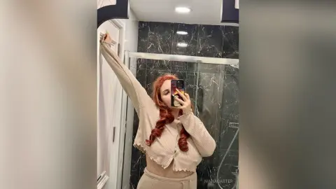 Redheaded woman in a beige ribbed cardigan taking a mirror selfie in a modern marble bathroom.