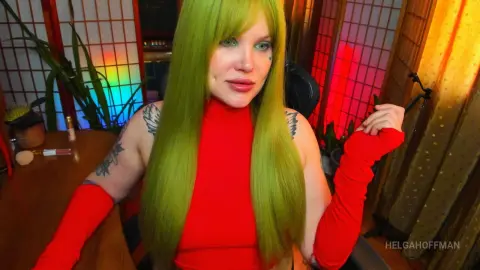 Woman with long lime green hair in a red outfit sitting in a room with colorful lighting.