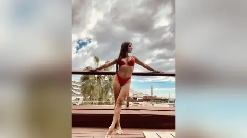 Rebecca poses in a red bikini on a balcony against a cloudy sky.