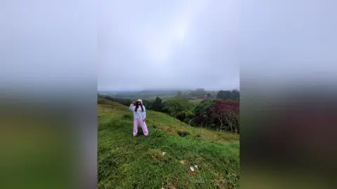 Woman in a white sweatshirt and pink pants standing on a lush green hillside under a cloudy sky.